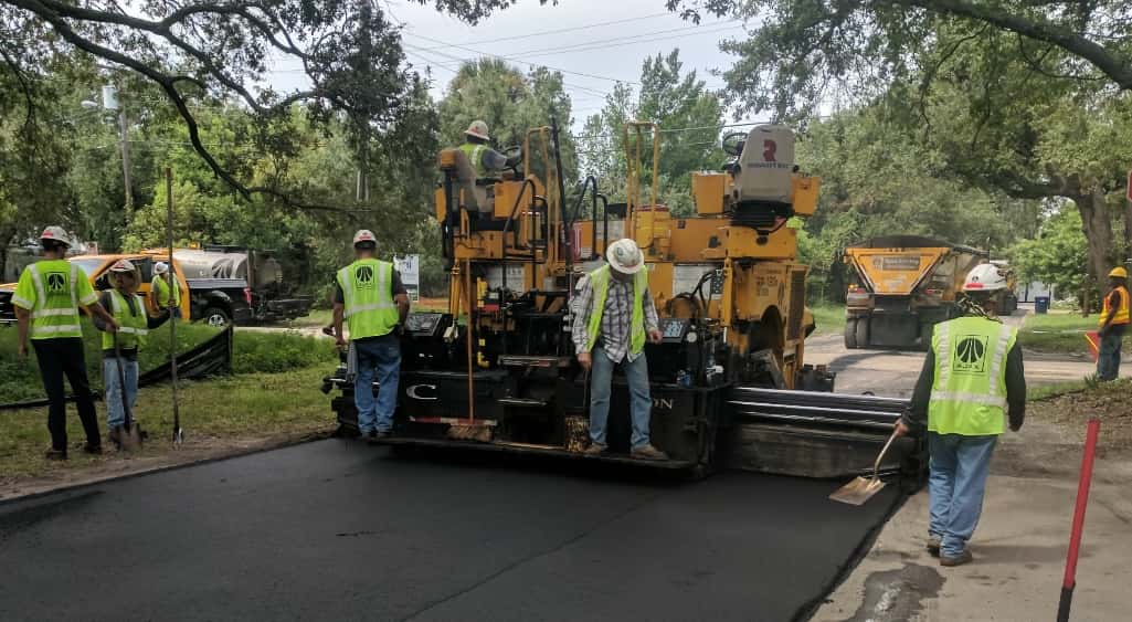 Heavy machinery used for resurfacing roads with a crew working in a neighborhood. Large oaks can be seen hanging over the road and in background.