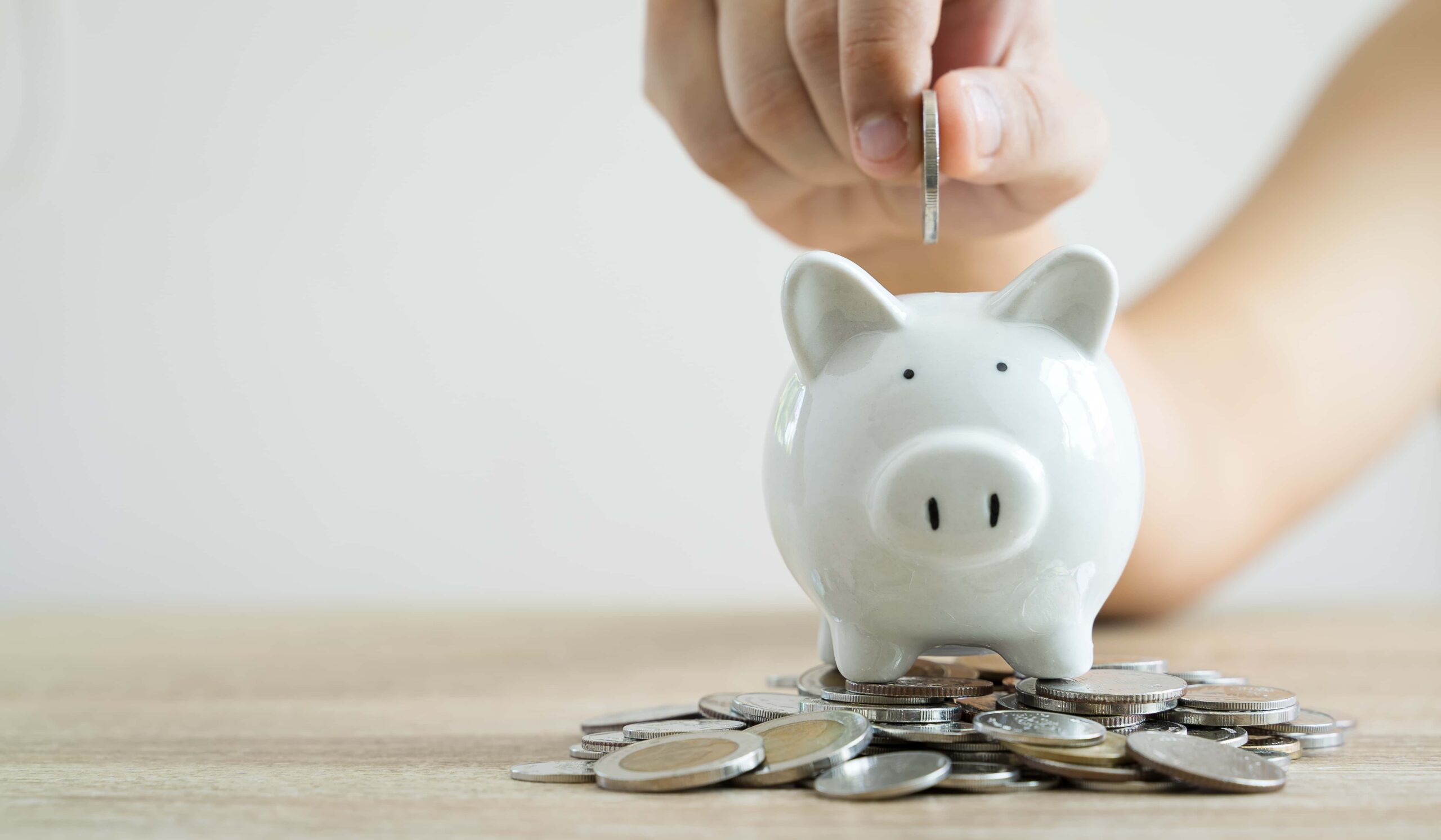 hand dropping coins into a small white piggy bank. More coins around bank on table.